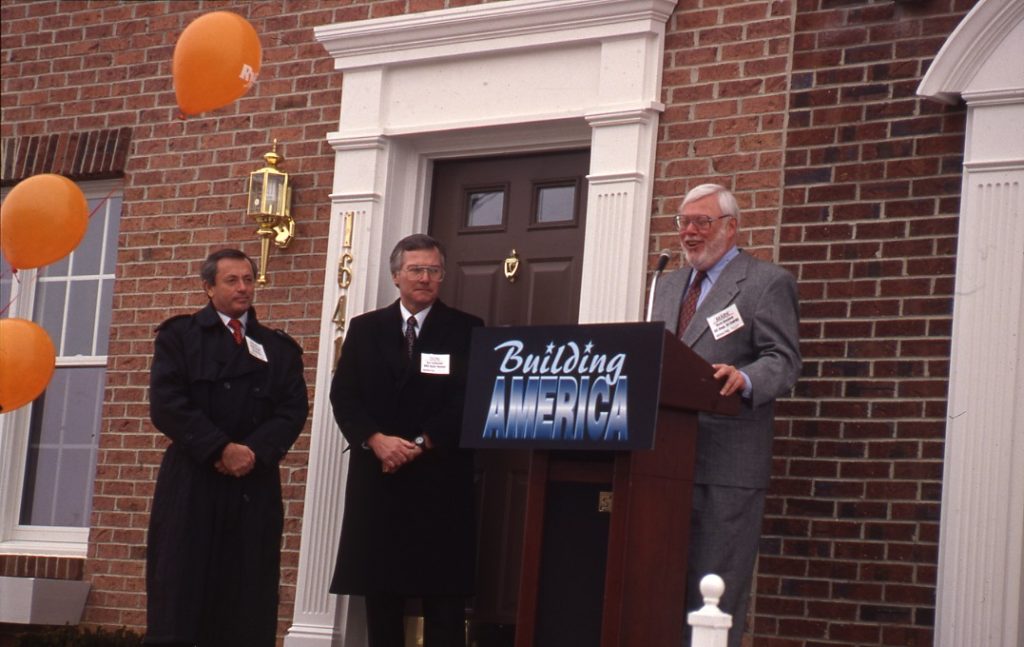 Three people, including Steven Winter, stand by a Building America podium in front of a building.