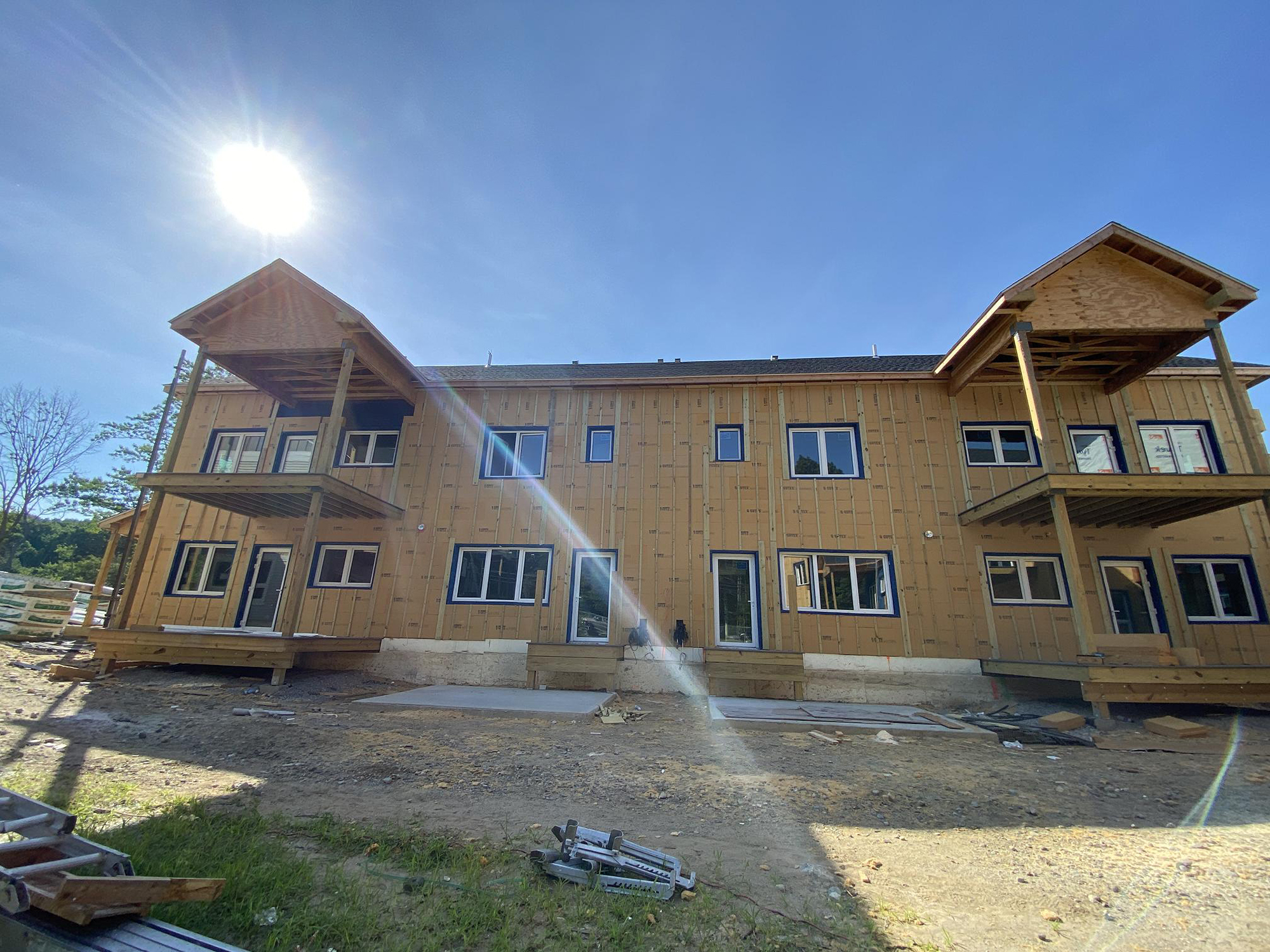 Partially constructed multi-unit residential building at Eagleville Green. The wooden exterior features multiple doors and windows across two levels, with covered balconies on each end. Construction materials and tools are visible in the foreground, and bright sunlight creates shadows and lens flare.
