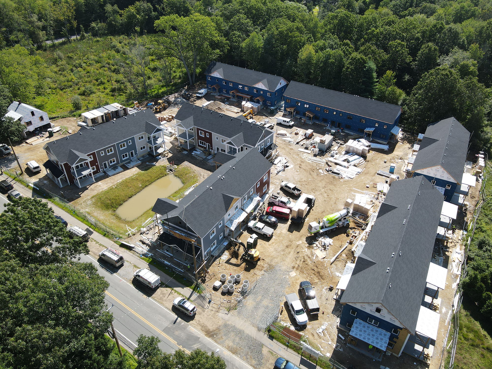 Aerial view of the Eagleville Green construction site showing multiple residential buildings in various stages of completion. Structures are arranged around a central area with construction materials, vehicles, and a small pond. The site is surrounded by wooded areas, highlighting its suburban setting.