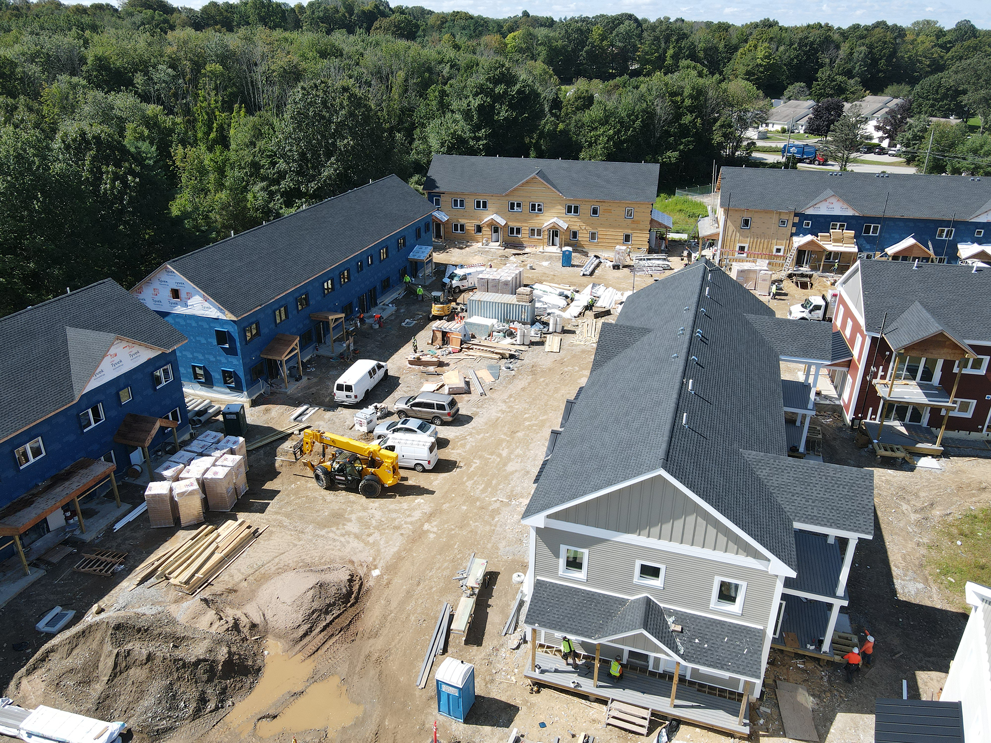 Construction site at Eagleville Green showing multiple buildings in various stages of completion. Some structures have blue or gray exteriors, while others display exposed wooden framing. A yellow construction vehicle, white vans, and piles of building materials are visible in the central dirt area. Trees and nearby residential buildings surround the site.