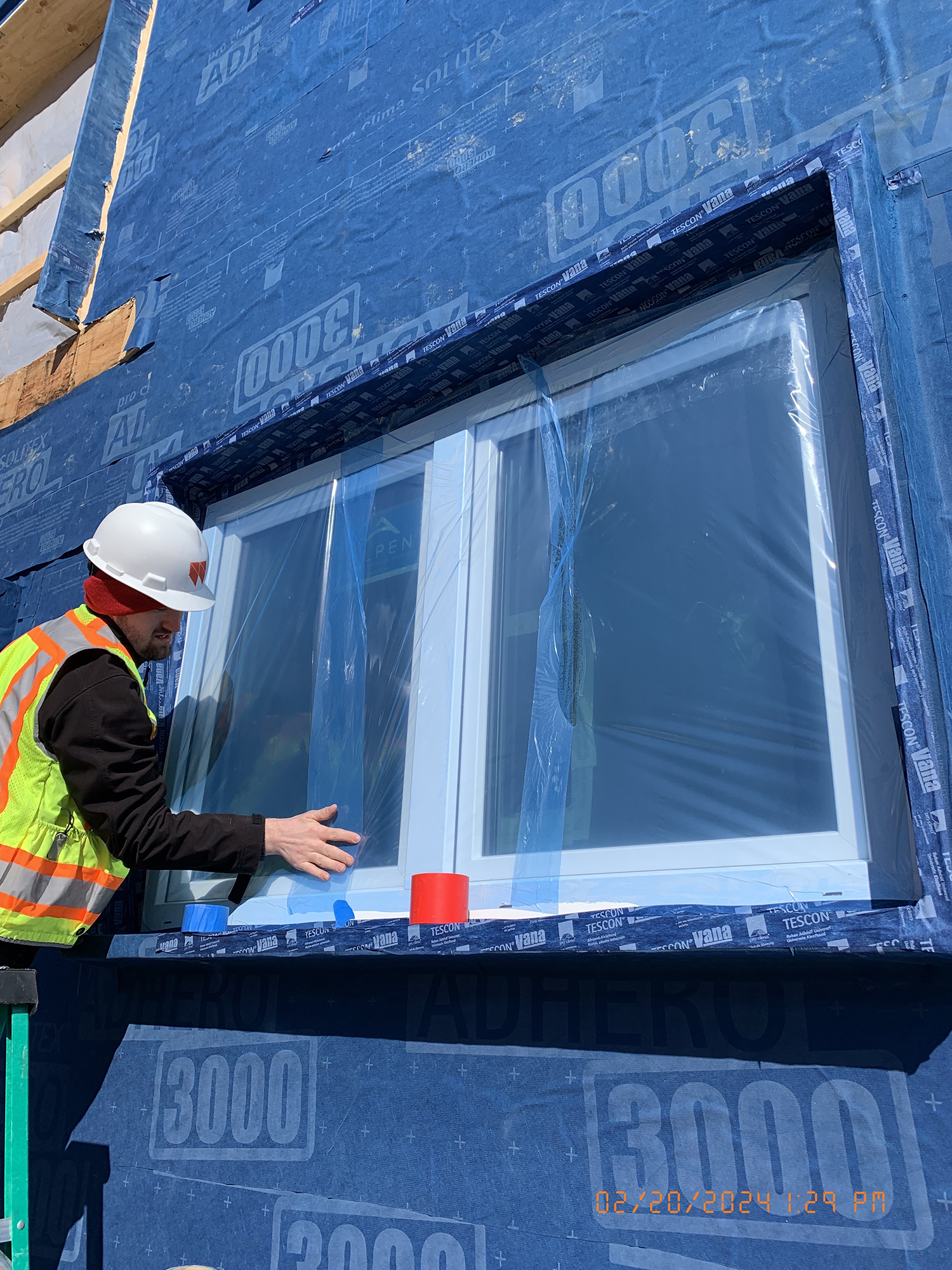 Person wearing a hard hat and high-visibility vest inspecting a window on a building exterior covered with blue weather-resistant barrier labeled '3000' and 'TESCON.'