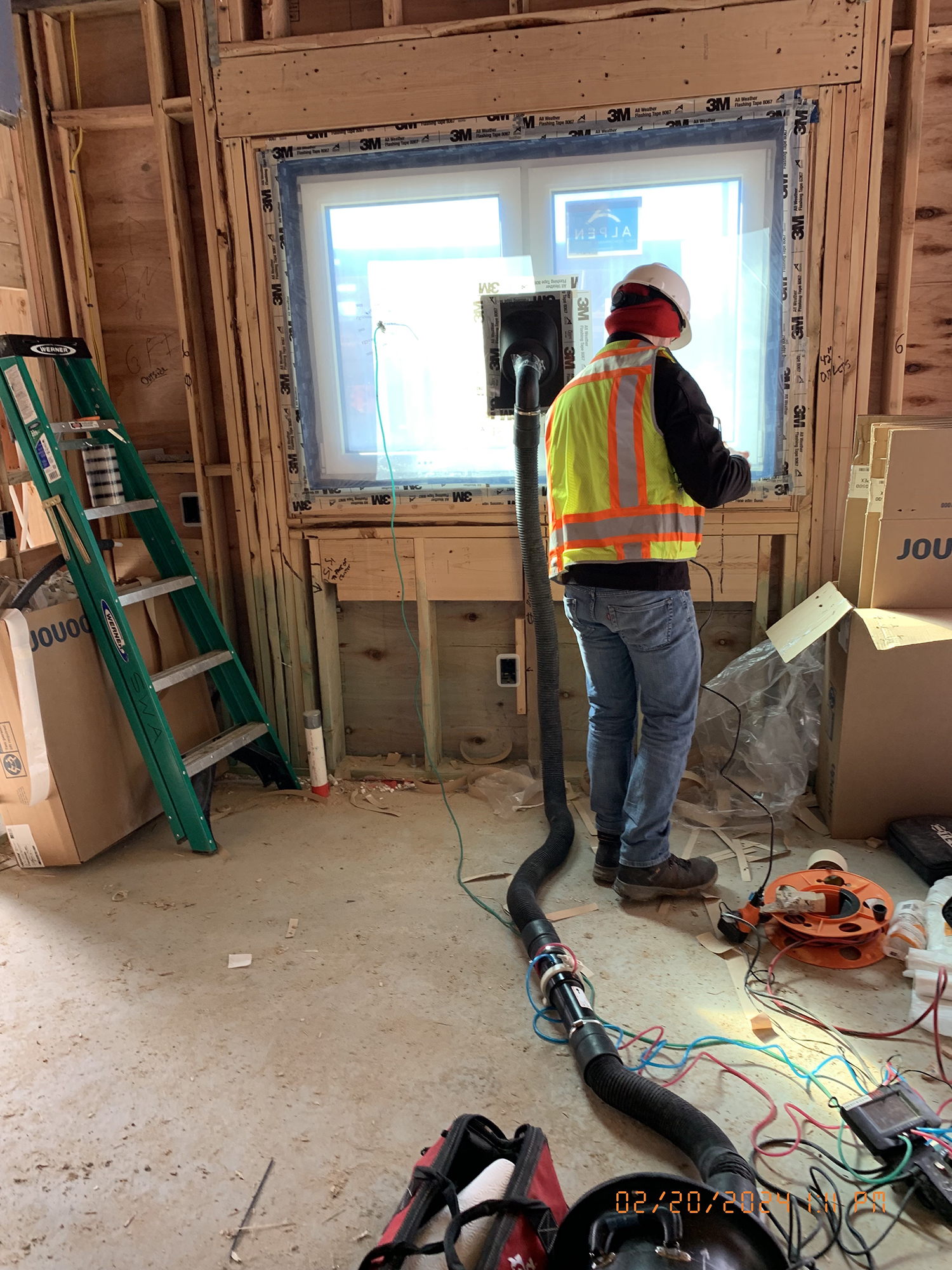Person wearing a safety vest and hard hat working near a window at the Eagleville Green site. Equipment is connected to the window, for testing. A green ladder, boxes labeled 'JOU,' and electrical cords are visible on the floor.