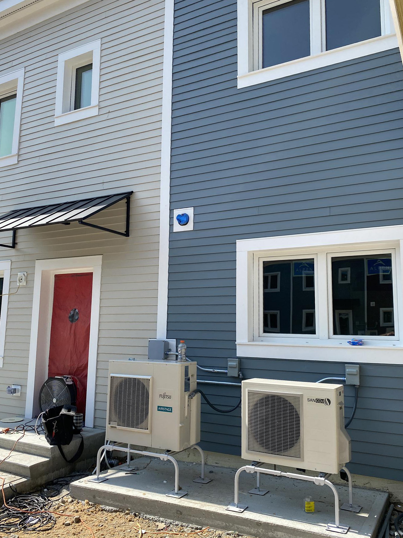 Exterior view of a building at Eagleville Green featuring light gray and blue siding. A red door with a black awning is on the left, with three windows above and beside two air conditioning units mounted on metal stands. Units are labeled 'Fujitsu Halcyon' and 'Samsung,' with electrical conduits and construction equipment visible near the ground.