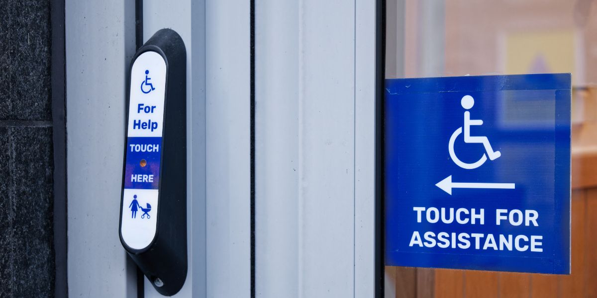 Accessibility assistance button mounted beside a building entrance door, next to a sign with the International Symbol of Access and the words "Touch for Assistance."