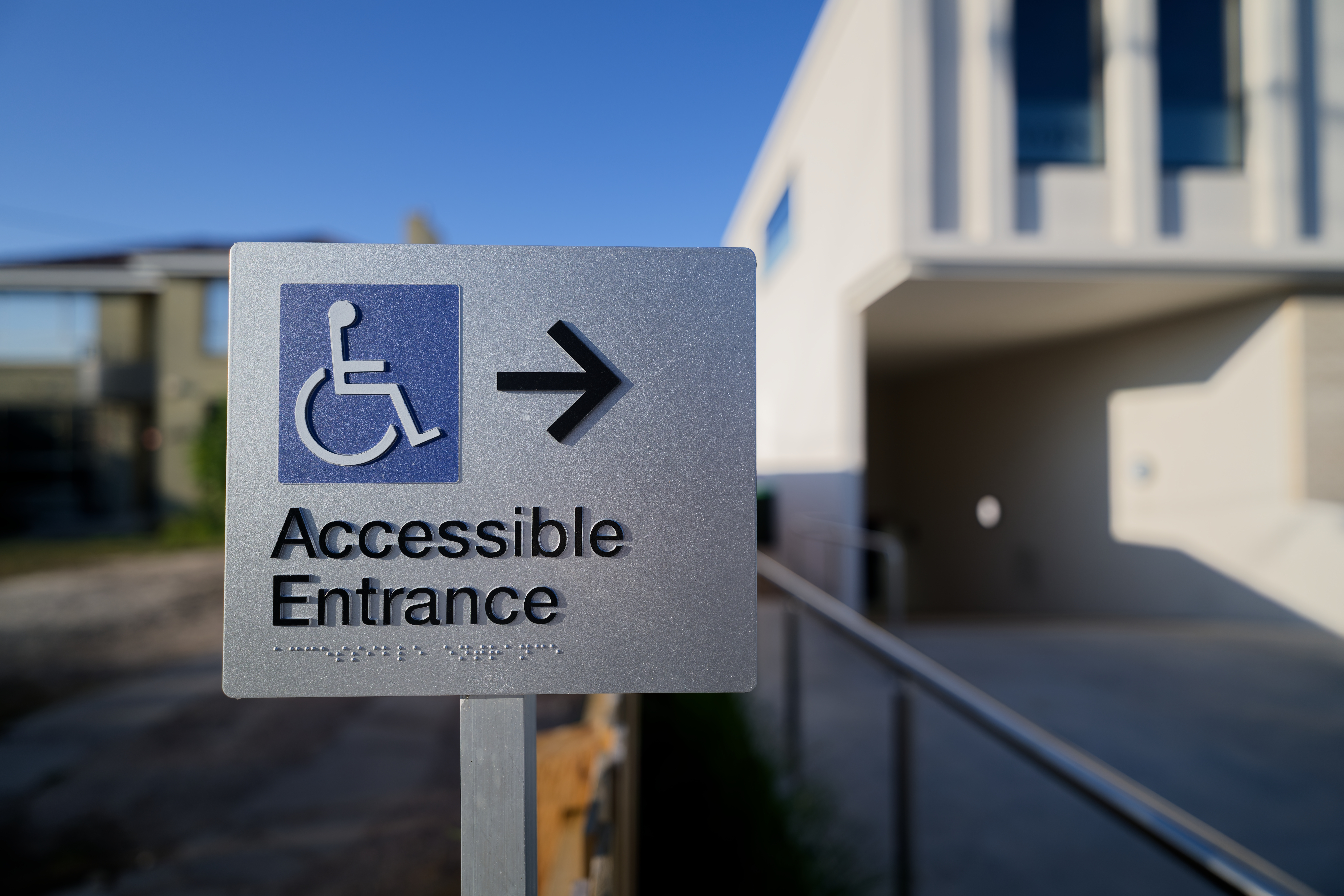 Accessible entrance sign with the International Symbol of Access and directional arrow, positioned in front of a building entrance and accessible route