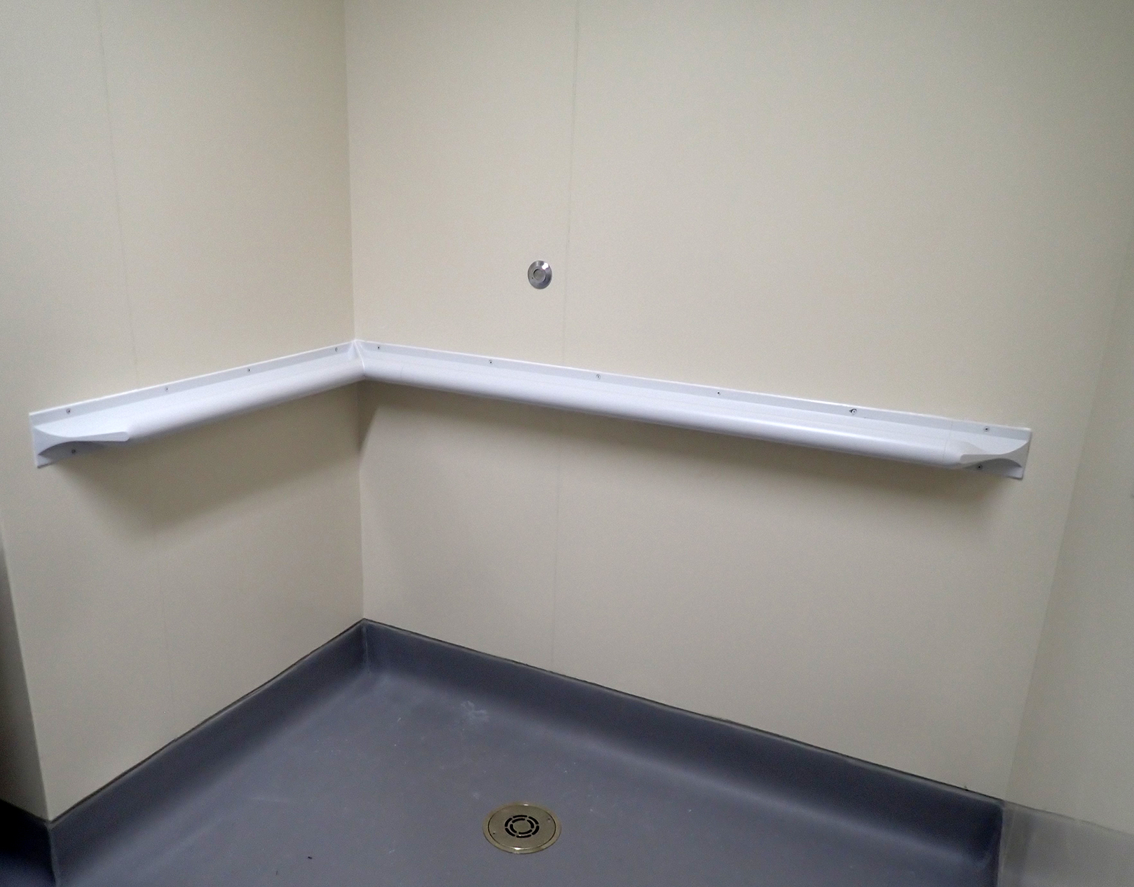 Photo of a white ligature-resistant grab bar installed in an L-shaped configuration in the corner of a shower. The grab bar is mounted horizontally on two adjacent beige walls and secured with multiple screws. The shower has a gray floor with a central drain.