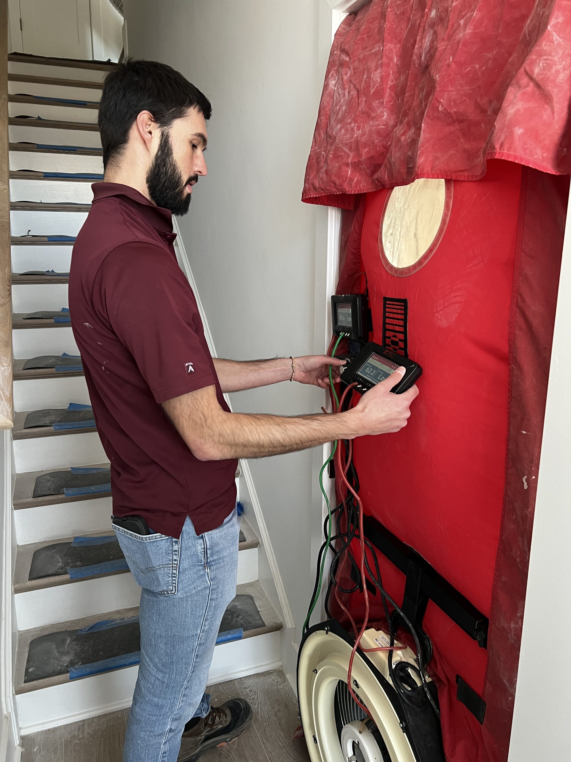 Person operating blower door testing equipment installed in a doorway, with the red blower door panel and pressure gauge visible and a staircase in the background.