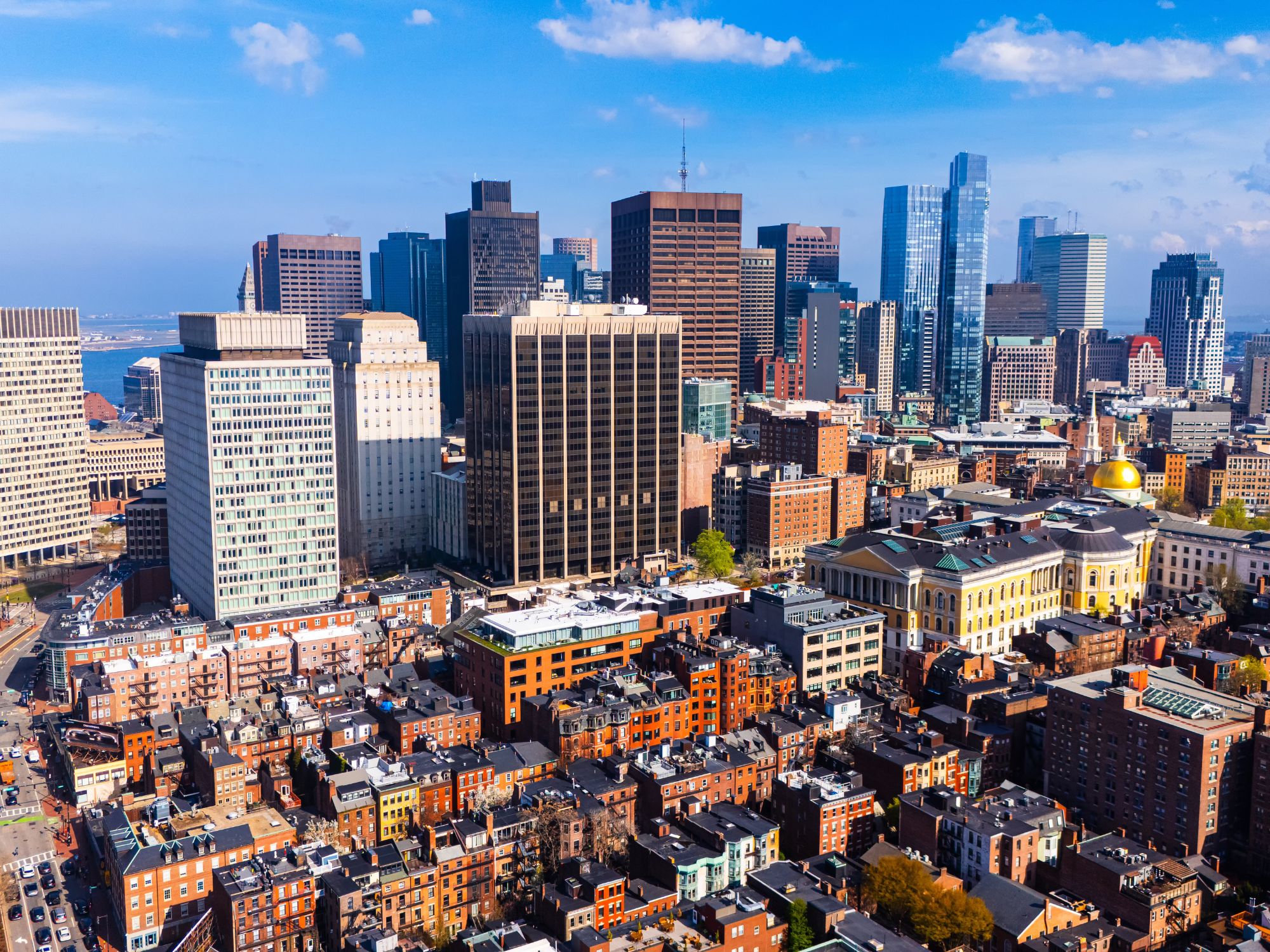 Aerial view of dense urban neighborhoods in Boston with mid-rise residential buildings in the foreground and a skyline of high-rise buildings in the background.