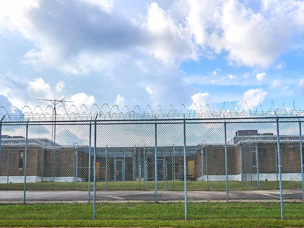 Single‑story detention center surrounded by a chain‑link fence topped with barbed wire, under a partly cloudy sky.
