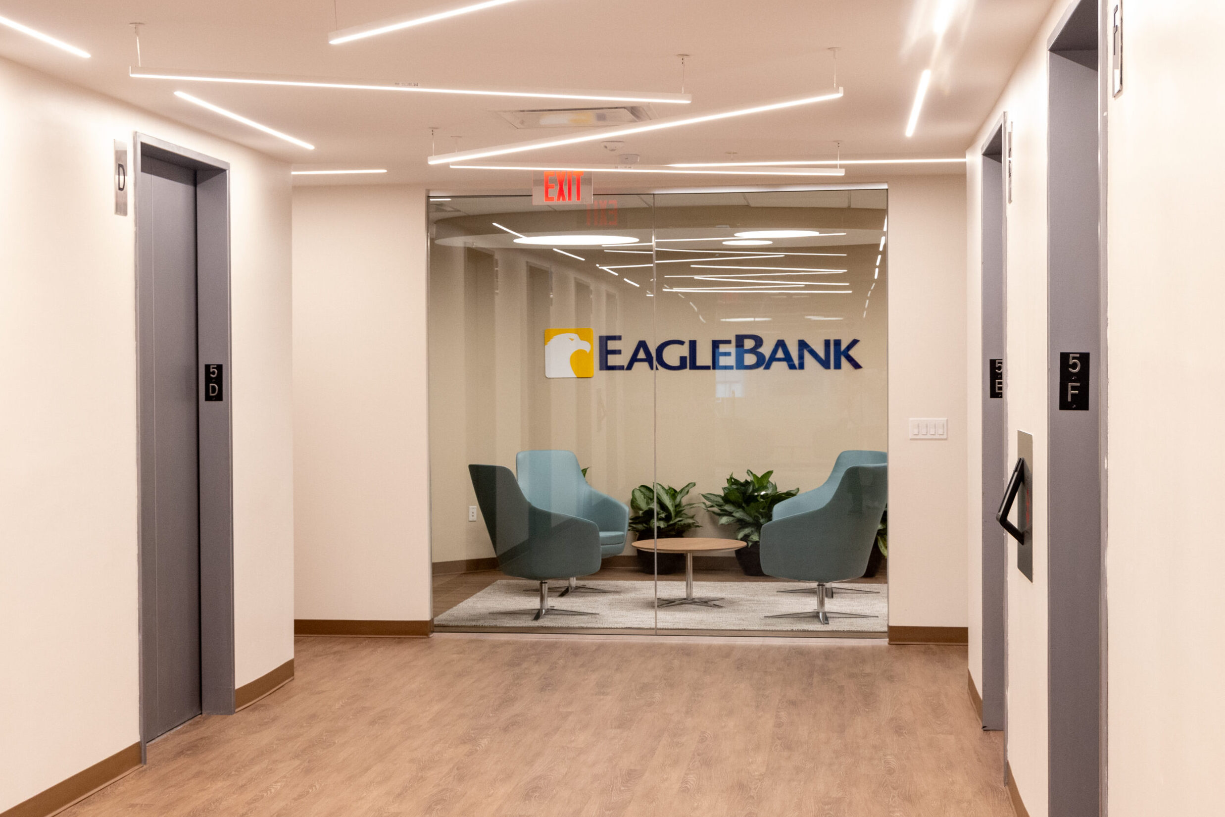 Lobby hallway leading to a glass-walled seating area with the EagleBank logo, featuring modern lighting, wood flooring, and two elevators on either side.