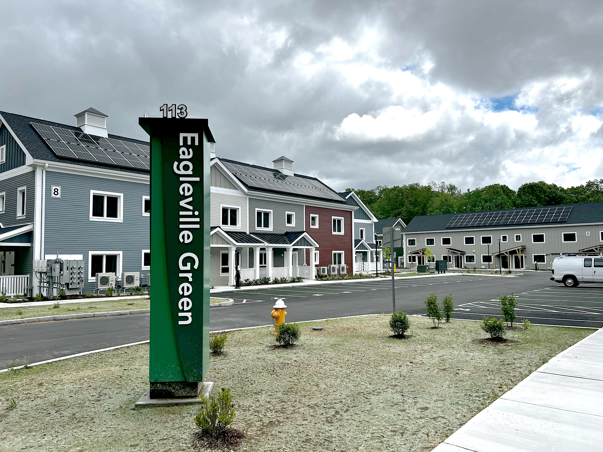 Entrance view of Eagleville Green featuring modern gray and red townhouses with rooftop solar panels. A green sign with white text displays the community name in the foreground, with small plants, a fire hydrant, and an overcast sky completing the scene.