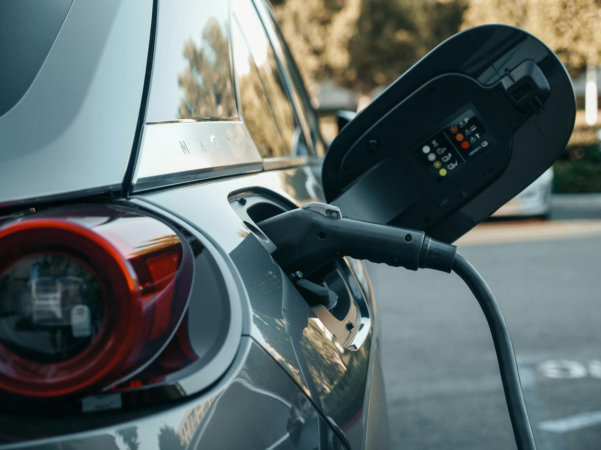 Photo of an electric car being charged. A charging cable is plugged into the car’s rear charging port, with the port cover open showing indicators and symbols. The car’s taillight and part of its body are also visible.