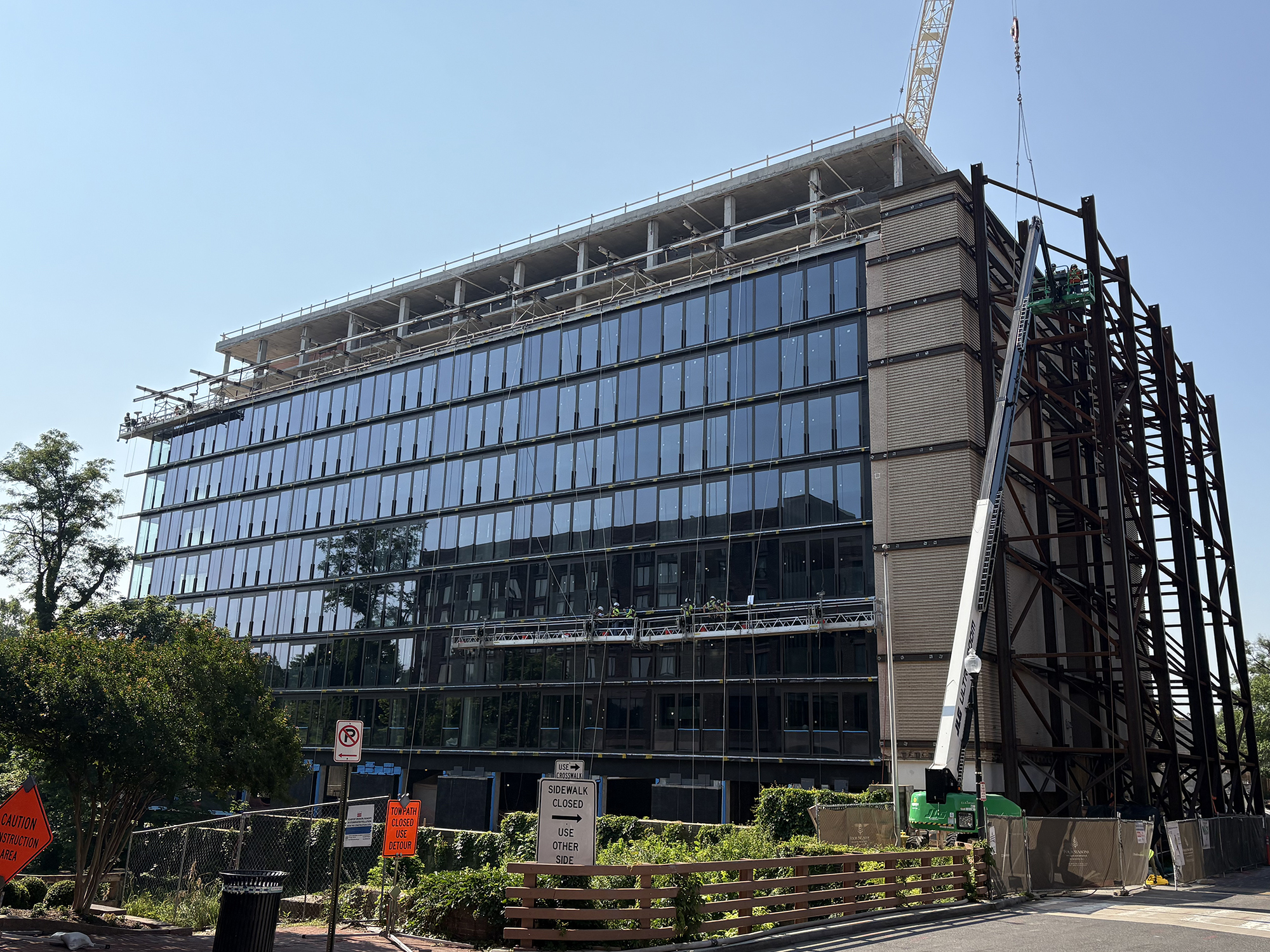 Four Seasons Private Residences under construction in Georgetown, Washington, DC. The modern multi-story building features extensive glass windows. A crane is positioned on the right, with construction signs, fencing, and greenery in the foreground. The site, formerly a coal yard, is being redeveloped into luxury housing with a one-acre public park.