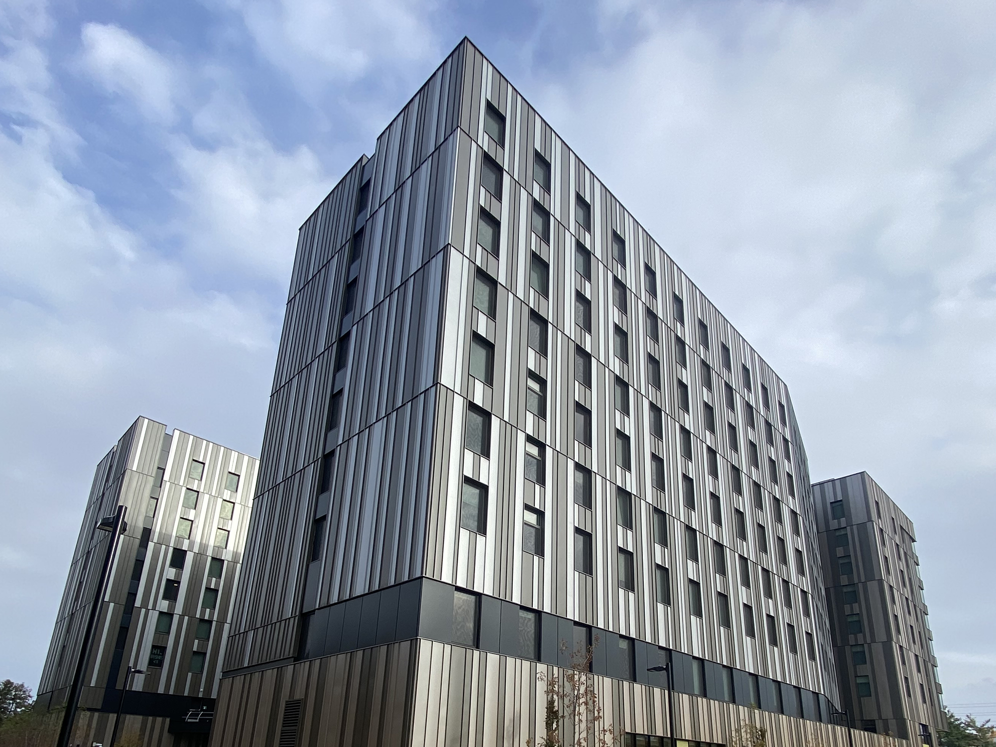 Exterior view of Harmony Commons student residence at the University of Toronto, featuring two modern mid-rise buildings with vertically striped metal cladding and rows of rectangular windows.