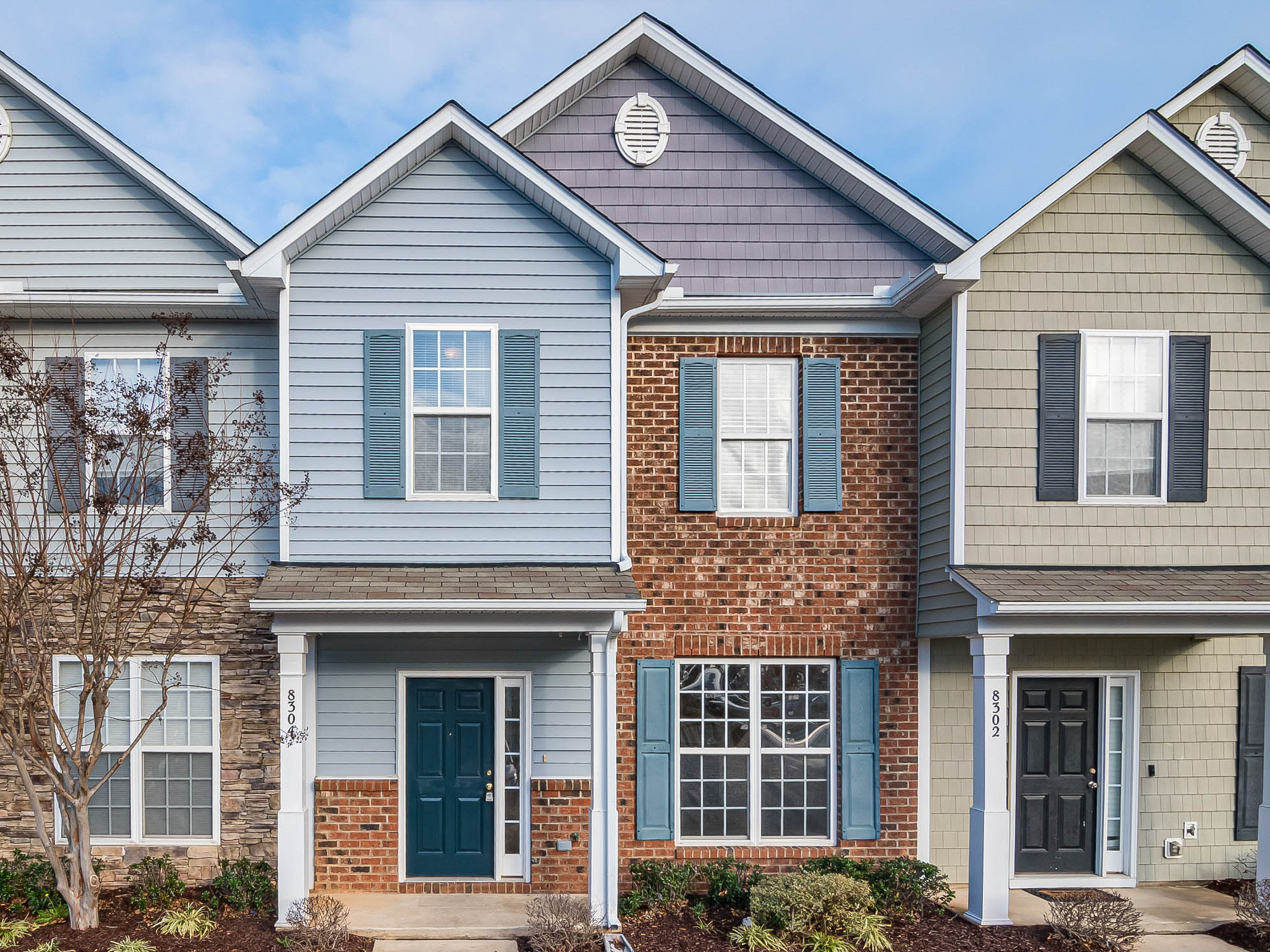 A row of three townhouses with distinct exteriors: one with light blue siding and blue shutters, one with a brick facade and blue shutters, and one with beige siding and black shutters. Each townhouse has a gabled roof with a circular vent near the peak, and a small tree is in front of the leftmost unit.