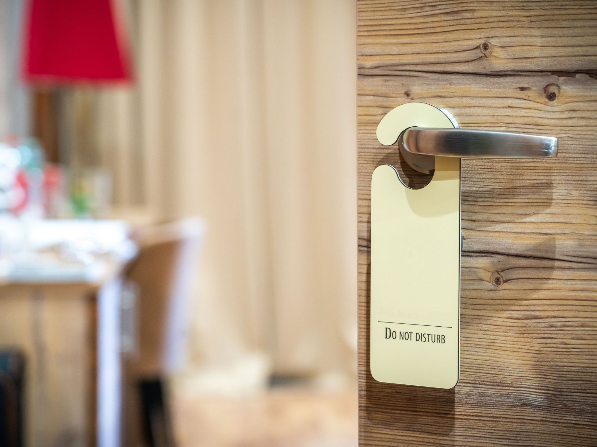 Hotel room door with a metal handle and a “Do Not Disturb” sign hanging on it; interior of the room with furniture and curtains visible in the background.
