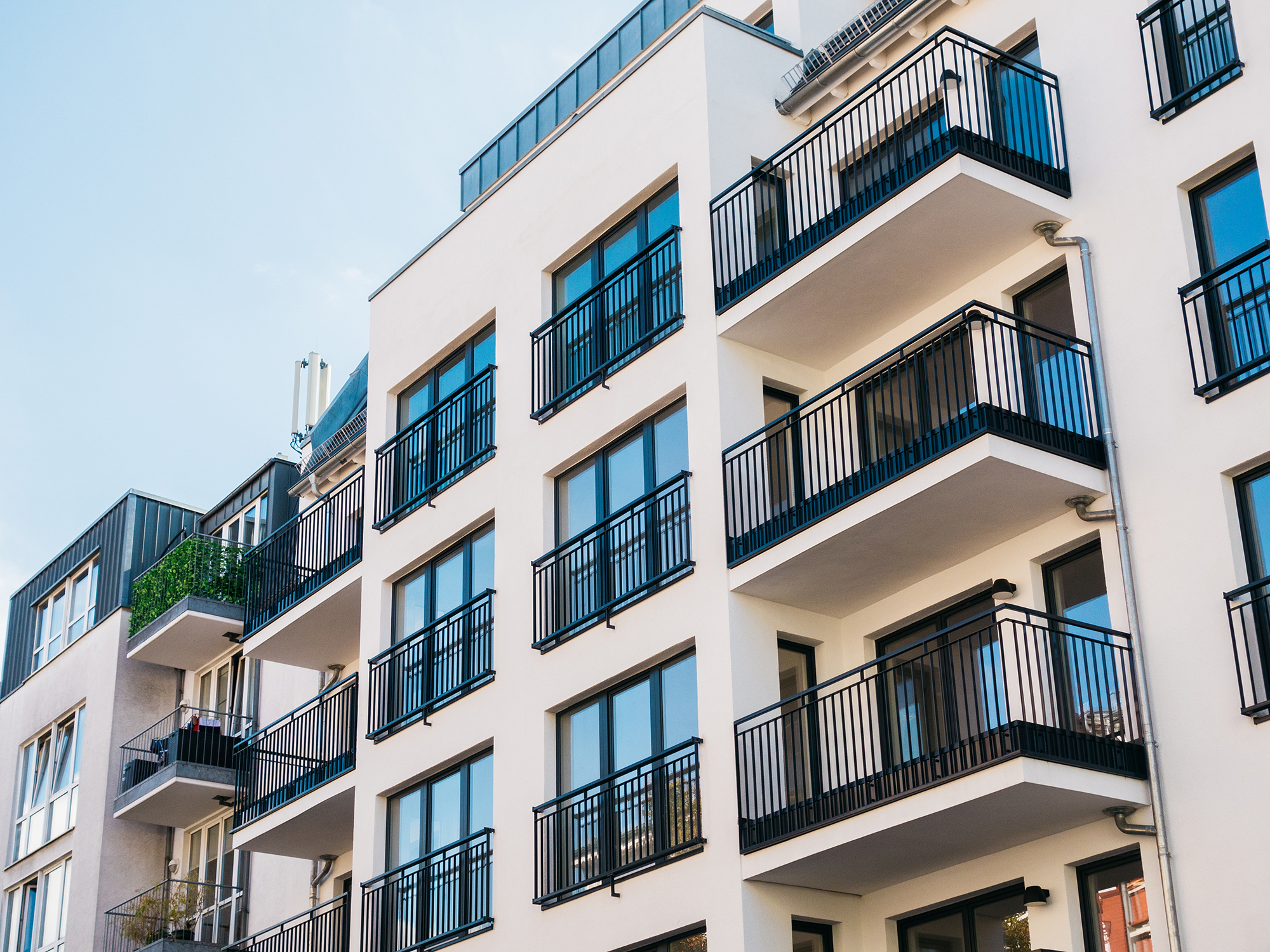 A modern apartment building with multiple floors, each featuring several balconies with black metal railings. The building’s facade is light-colored, and the windows are large, allowing for ample natural light. The architectural design is contemporary, with clean lines and a minimalist aesthetic.