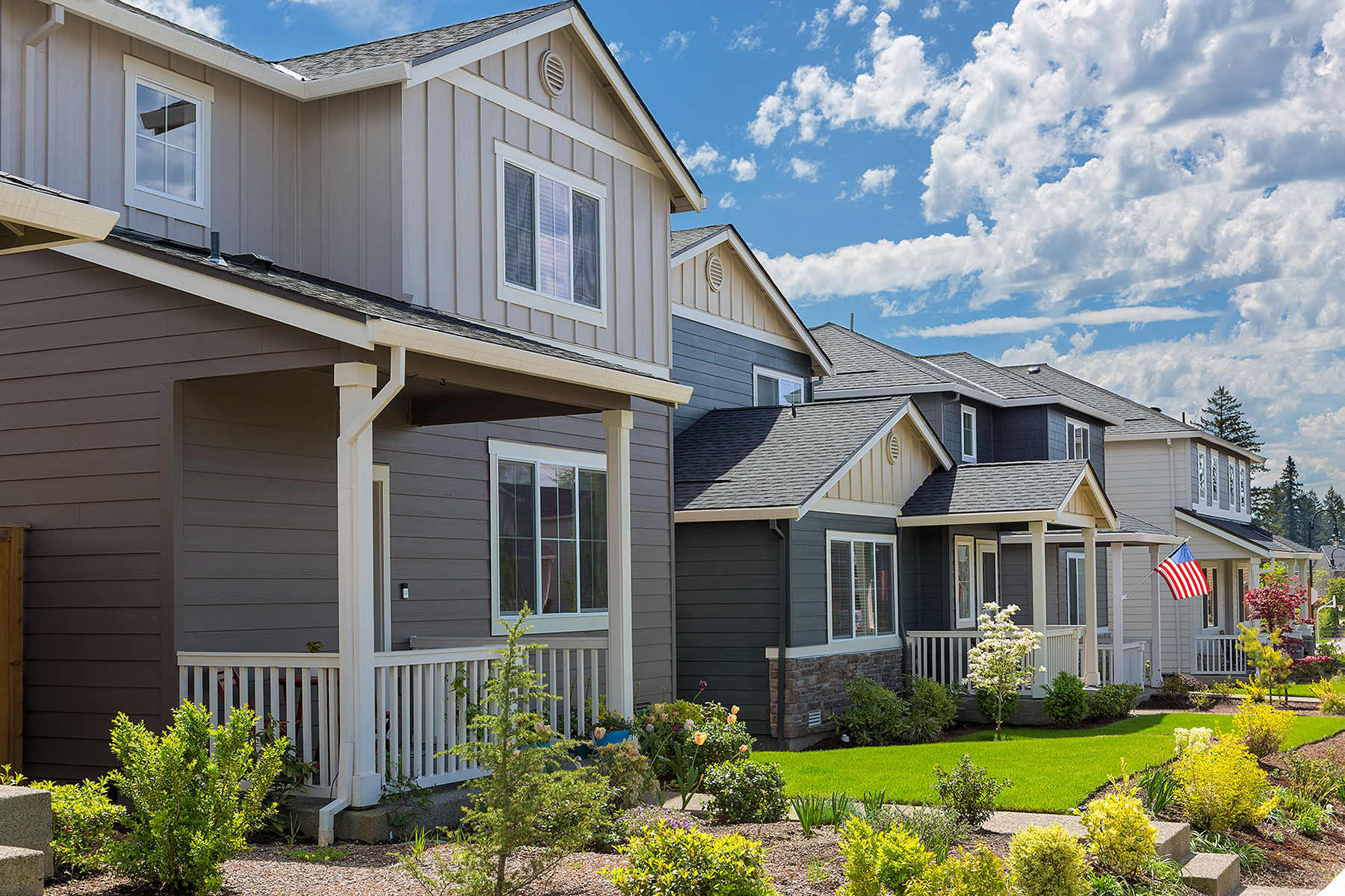 Row of newly built single-family homes with front porches, landscaped yards, and sidewalks in a suburban neighborhood under a bright blue sky.