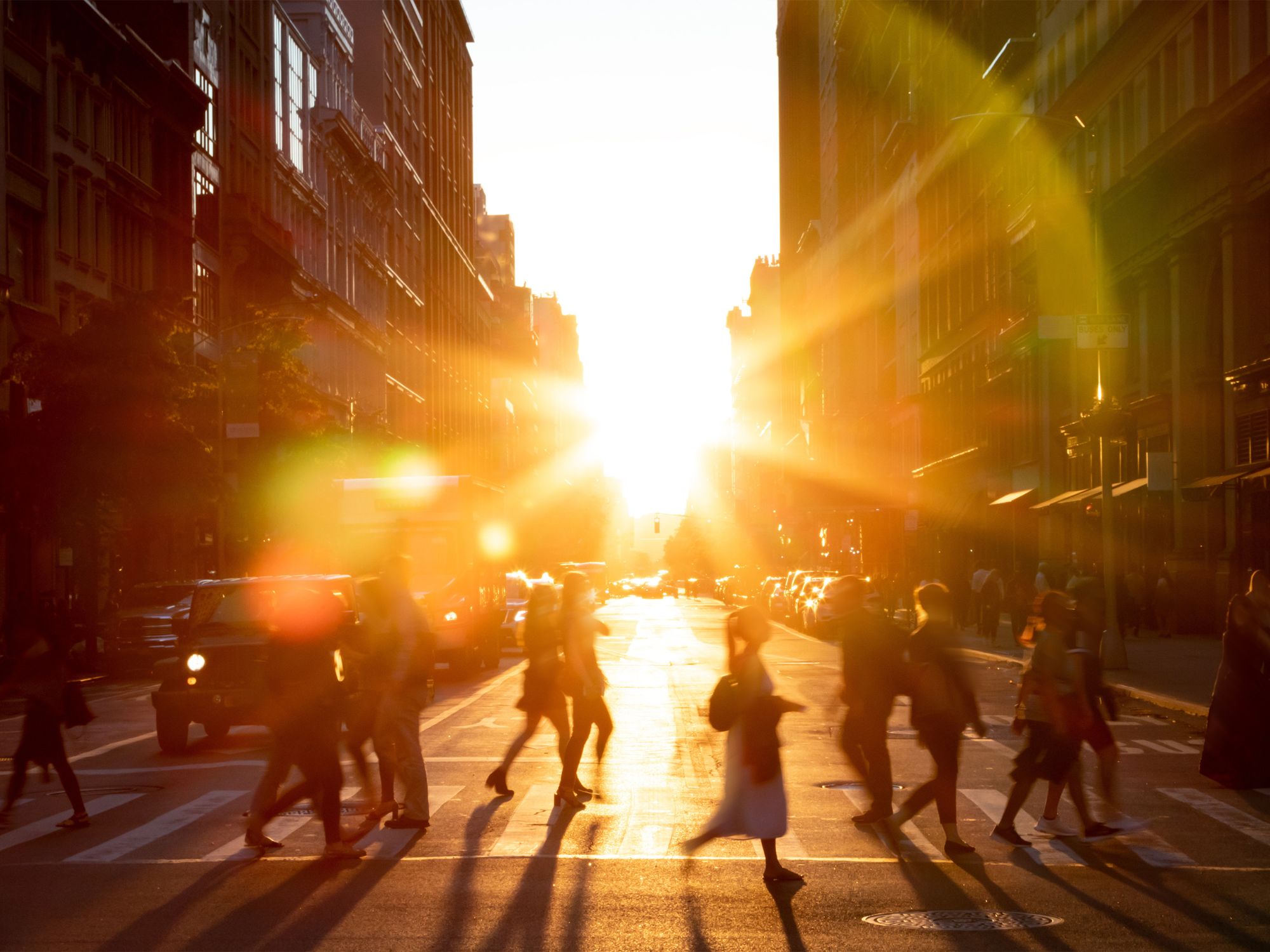 Sunlit city street with pedestrians crossing and strong heat haze between tall buildings.