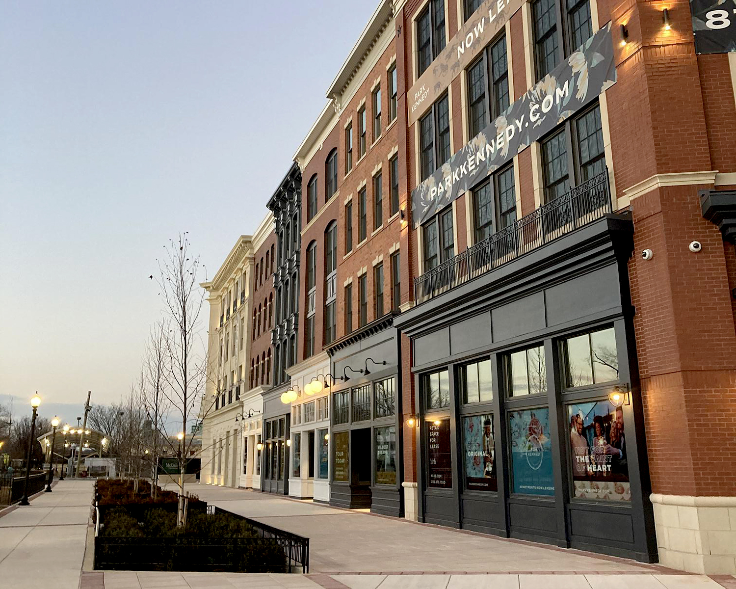 A view of the Park Kennedy development in Hill East, Washington, DC, showing newly constructed buildings with brick and light-colored facades, large windows, and retail spaces. A banner reading "ParkKennedy.com" promotes leasing. The wide sidewalk is lined with young trees and street lamps under a clear sky.