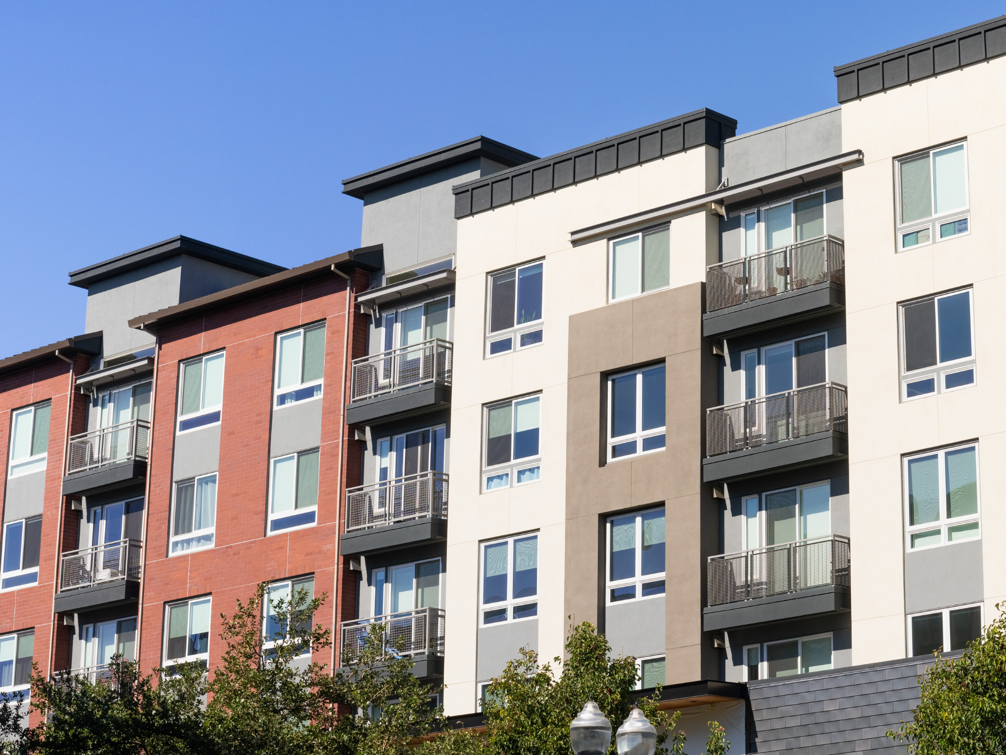 Modern mid-rise apartment building with mixed exterior finishes, including red brick and neutral-colored panels, featuring multiple balconies and large windows.