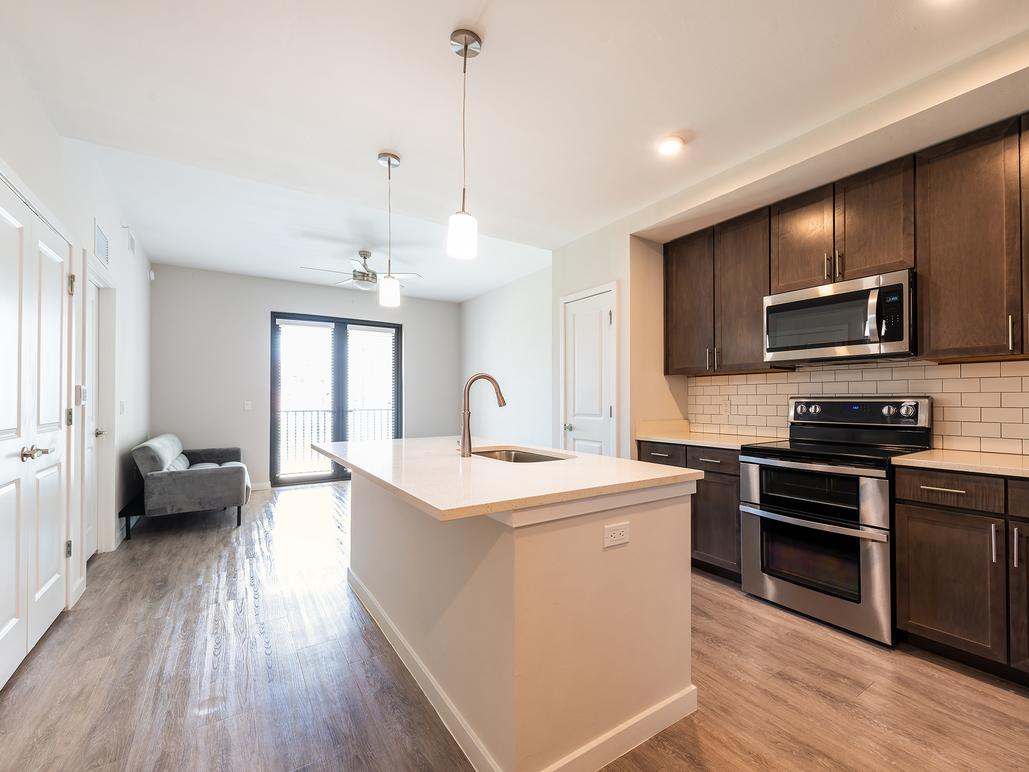 A modern open-concept kitchen and living area featuring dark wooden cabinets, stainless steel appliances, a white subway tile backsplash, and a kitchen island with a white countertop and sink.