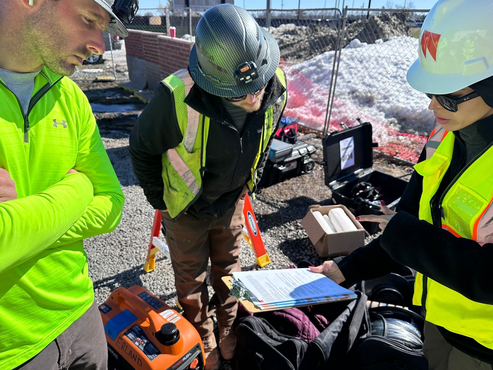 Construction workers wearing safety helmets and high-visibility vests review equipment and materials on an active job site next to a partially built structure