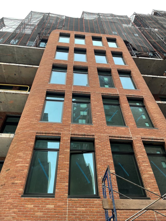 Mid-rise brick façade with grid of windows and rounded corner, shown during construction with scaffolding above