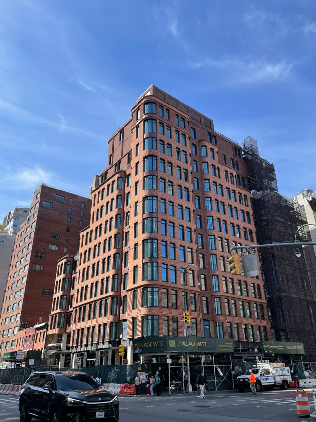 Completed brick residential building at street corner with rounded edges, ground-floor storefronts, and surrounding city context
