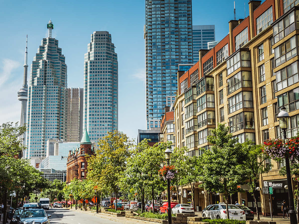 A vibrant Toronto street scene featuring a mix of modern skyscrapers and historic buildings. The CN Tower is visible in the background, while a historic flatiron building with a green roof stands in the foreground surrounded by trees.
